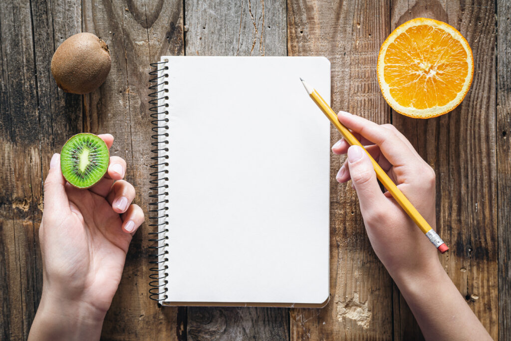 Woman's hands with a notebook, holding a pencil and a kiwi, there is also half of an orange.